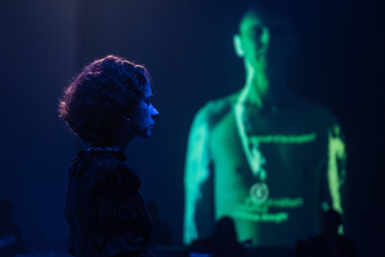 woman standind in front of dark blue background, green video projection in the background